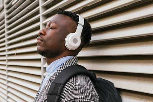 Seitenansicht eines jungen, aufmerksamen afroamerikanischen männlichen Büroangestellten, der mit geschlossenen Augen über ein drahtloses Headset in der Nähe einer gerippten Wand in der Stadt ein Lied hört - ADSF22806