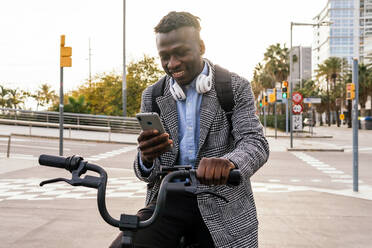 Young glad black male office worker with headset chatting on cellphone while sitting on bike on urban pavement - ADSF22808