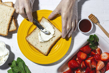 Oben Ansicht Ernte anonyme weibliche Koch in Latex-Handschuhen Verbreitung leckeren Frischkäse auf Brotscheibe auf gelben Teller neben geschnittenen Erdbeeren platziert - ADSF22822