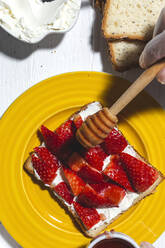 Overhead Ernte anonym qualifizierte weibliche Köchin in Handschuhen gießen süßen Honig auf leckeren Toast mit Frischkäse und schneiden Erdbeeren beim Kochen in hellen Küche - ADSF22824
