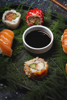 Leckere, frisch zubereitete Sushi auf grünen Pflanzenzweigen auf einem schwarzen Teller mit Sojasauce auf einem Marmortisch in der Nähe von Stäbchen - ADSF22825