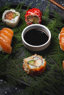 Tasty fresh assorted sushi served on green plant twigs on black plate with soy sauce on marble table near chopsticks - ADSF22825