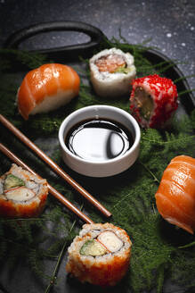 Leckere, frisch zubereitete Sushi auf grünen Pflanzenzweigen auf einem schwarzen Teller mit Sojasauce auf einem Marmortisch in der Nähe von Stäbchen - ADSF22826