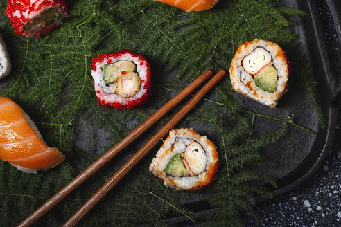 Leckere, frisch zubereitete Sushi auf grünen Pflanzenzweigen auf einem schwarzen Teller mit Sojasauce auf einem Marmortisch in der Nähe von Stäbchen - ADSF22827