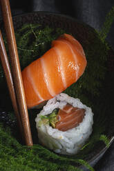Leckere, frisch zubereitete Sushi auf grünen Pflanzenzweigen auf einem schwarzen Teller mit Sojasauce auf einem Marmortisch in der Nähe von Stäbchen - ADSF22828