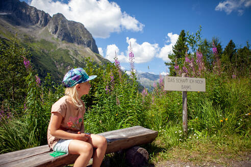 Girl looking at sign board while sitting on bench during sunny day - GAF00190