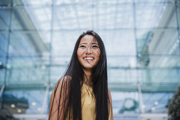 Smiling woman looking away while standing in front of modern building - EBBF03335