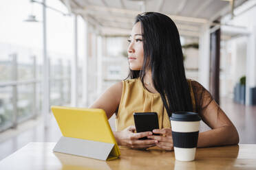 Unternehmerin schaut weg, während sie ihr Smartphone in einer modernen Büro-Cafeteria hält - EBBF03348