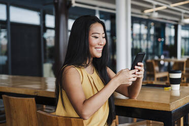 Lächelnde weibliche Fachkraft, die ein Smartphone benutzt, während sie in einer Büro-Cafeteria sitzt - EBBF03356