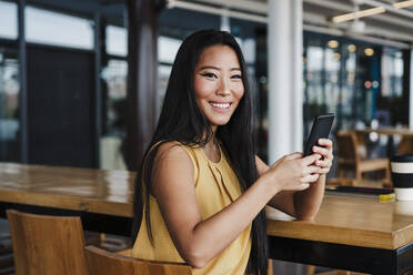 Eine Unternehmerin hält ein Mobiltelefon in der Hand und sitzt auf einem Stuhl in einem modernen Büro - EBBF03357