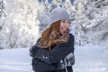 Lächelnde junge Frau, die sich selbst umarmt, während sie im Winter wegschaut - LBF03507