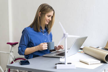 Geschäftsfrau hält Kaffeetasse und benutzt Laptop im Büro - GIOF12448