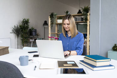 Smiling businesswoman with books on desk using laptop at office - GIOF12479