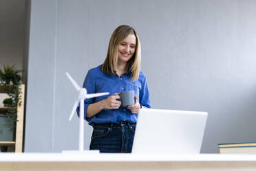 Smiling businesswoman holding coffee cup while looking at laptop in office - GIOF12493