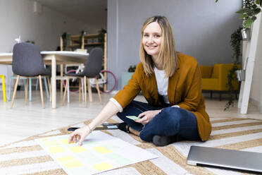 Smiling creative businesswoman with adhesive notes sitting on carpet in office - GIOF12498