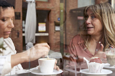 Female friends chatting while having coffee at cafeteria seen through glass - VEGF04213