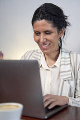 Lächelnde weibliche Fachkraft, die in der Cafeteria am Laptop arbeitet - VEGF04237