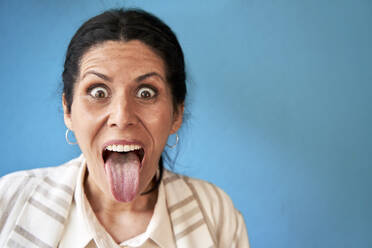 Reife Frau streckt die Zunge vor einer blauen Wand heraus - VEGF04244