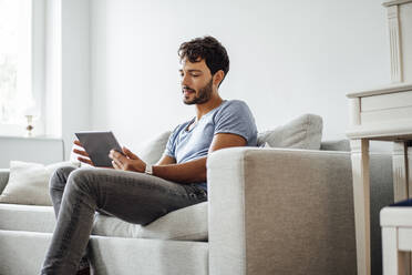 Young man using digital tablet while sitting on sofa in living room - JOSEF04030