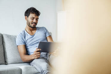 Smiling young man using digital tablet while sitting on sofa at home - JOSEF04031