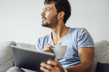 Junger Mann sitzt mit Kaffeetasse und Tablet und schaut weg - JOSEF04033