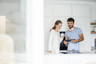Smiling young couple discussing over digital tablet while standing in kitchen - JOSEF04036
