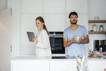 Smiling young woman using tablet while man listening music in kitchen - JOSEF04037