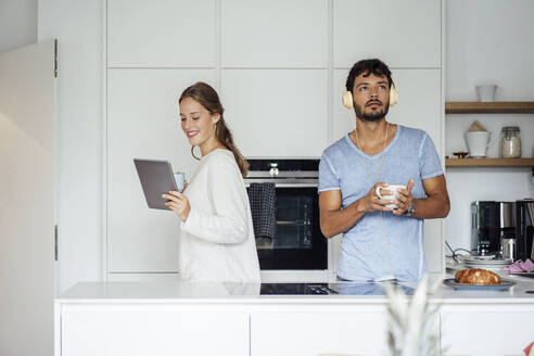 Lächelnde junge Frau, die ein Tablet benutzt, während ein Mann in der Küche Musik hört - JOSEF04037