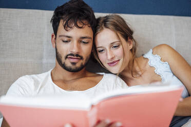 Young couple reading book while sitting on bed at home - JOSEF04064