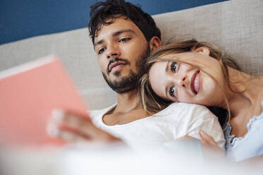 Young man with smiling woman reading book in bedroom - JOSEF04066