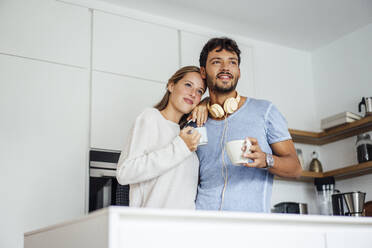 Smiling young couple holding coffee cup in kitchen while looking away - JOSEF04073