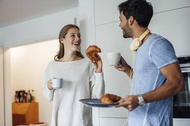 Happy young couple with croissant and coffee cup in kitchen - JOSEF04074