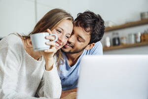 Happy man embracing girlfriend looking at laptop in kitchen - JOSEF04079