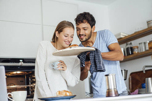 Mann zeigt seiner Freundin ein frisch gebackenes Croissant in der Küche zu Hause - JOSEF04080