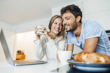 Happy young couple having coffee while leaning on kitchen counter at home - JOSEF04086