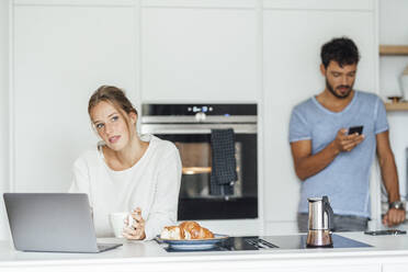 Thoughtful woman with laptop while boyfriend using smart phone in background at home - JOSEF04089