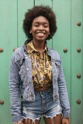 Curly hair woman smiling while standing in front of wall - JRVF00430