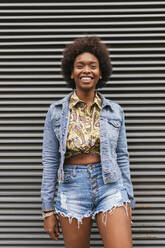 Smiling curly hair woman in front of corrugated wall - JRVF00432