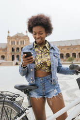 Woman using mobile phone while standing with bicycle on footpath - JRVF00441