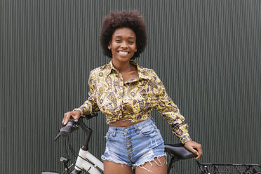 Smiling woman with bicycle standing in front of wall - JRVF00444