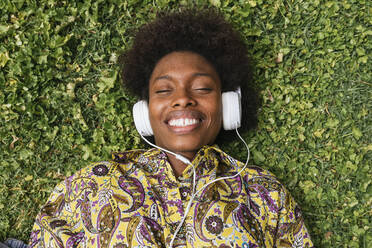 Smiling woman listening music through headphones while lying on grass - JRVF00449