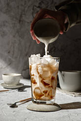 Anonyme Person gießt Milch aus einer Tasse in ein Glas mit Eiskaffee, das auf einem Tisch im Sonnenlicht steht - ADSF22845