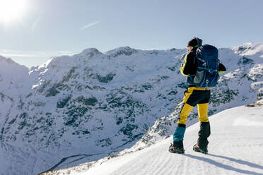 Full body back view of unrecognizable male mountaineer in warm activewear with backpack standing on slope of snowy rocky mountain and enjoying spectacular landscape in sunny winter day - ADSF22888