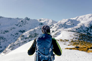 Back view of unrecognizable male mountaineer in warm activewear with backpack standing on slope of snowy rocky mountain and enjoying spectacular landscape in sunny winter day - ADSF22889