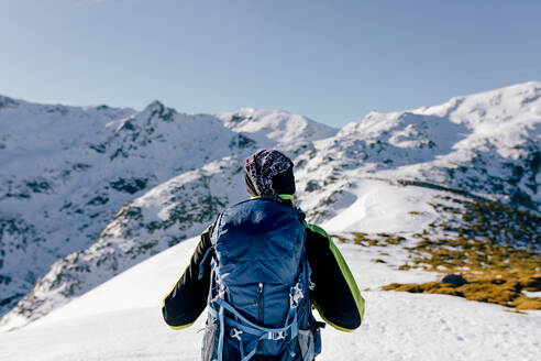 Rückenansicht eines nicht erkennbaren männlichen Bergsteigers in warmer Aktivkleidung mit Rucksack, der am Hang eines verschneiten felsigen Berges steht und die spektakuläre Landschaft an einem sonnigen Wintertag genießt - ADSF22889