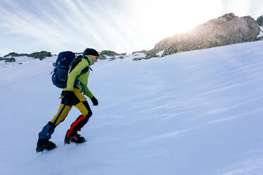 Side view of climber walking on slope of snow covered rocky mountain range in sunny weather - ADSF22890