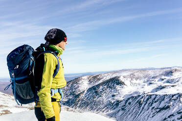 Side view of unrecognizable male mountaineer in warm activewear with backpack standing on slope of snowy rocky mountain and enjoying spectacular landscape in sunny winter day - ADSF22892