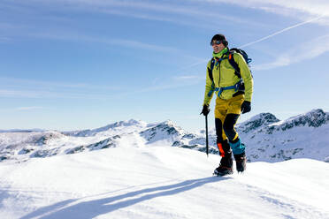 Male climber in outwear walking on slope of snow covered rocky mountain range in sunny weather - ADSF22894