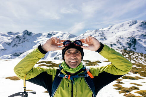 Selbstbewusster, glücklicher, männlicher Bergsteiger in stilvoller, warmer Aktivkleidung mit Rucksack und Kletterausrüstung, der auf dem Gipfel eines Berges steht und in die Kamera schaut, während er eine verschneite Felslandschaft bei sonnigem Wetter erkundet - ADSF22895