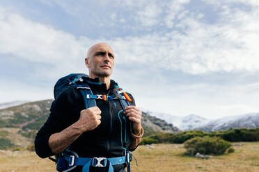 Erwachsener glatzköpfiger aktiver männlicher Bergsteiger in Aktivkleidung mit Rucksack, der sich auf einem Berggipfel ausruht und die malerische Aussicht auf eine schneebedeckte Bergkette genießt - ADSF22897